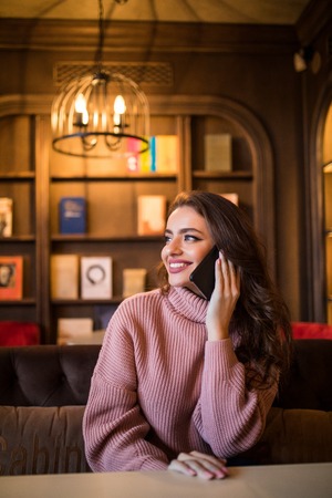 Young happy hipster girl speaking with her friend by mobile phone while relaxing in cozy coffee shopの写真素材