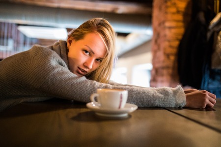 Beautiful young woman lies on hands, sits at wooden table in cafeteria, drinks coffee. Relax and rest conceptの写真素材