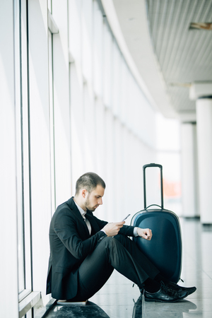 Young business man sitting on the phone with the suitcase at the airport waiting for the flightの写真素材