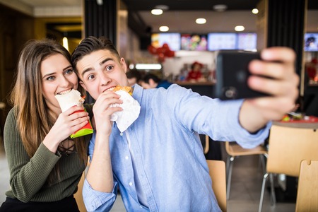 Happy couple eating burger and doner taking selfie together in fast foodの写真素材