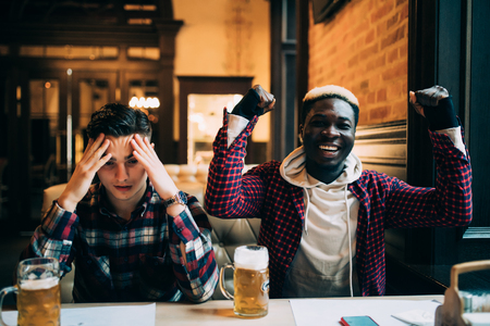 Afro american man happy shout his team score while other man upset. Men friends drinking beer and support different teams in the barの写真素材