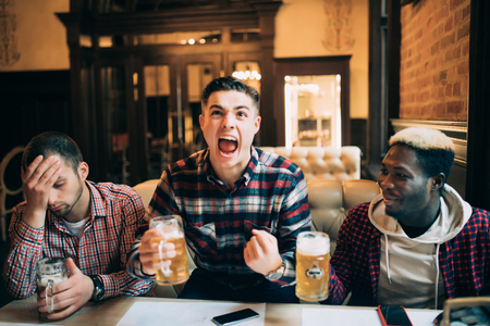Watching TV in bar. Two happy young men drinking beer and gesturing while sitting in barの写真素材