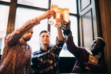 Multiracial friends men cheers with beer in glasses in pub.の写真素材