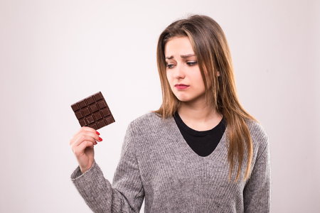 Happy young beautiful lady eating chocolate and smiling. Girl tasting sweet chocolate. Young woman with natural make up having fun and eating chocolate isolated on gray backgroundの写真素材