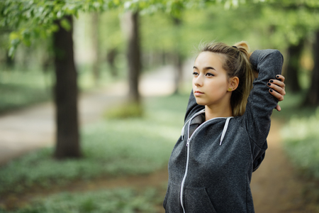 Closeup portrait of content young attractive woman wearing sportswear, raising clasped hands and stretching body in park with green view in background.の写真素材