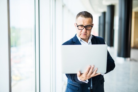 Cheerful mature man in formalwear adjusting his eyeglasses and holding laptop while standing in officeの写真素材