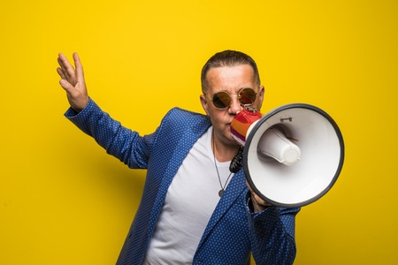 Mature man in sunglasses speaking with loudspeaker isolated on yellow backgroundの写真素材