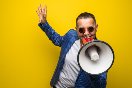 Mature businessman in sunglasses speaking with loudspeaker isolated on yellow backgroundの写真素材