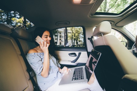 Businesswoman sitting on back seat of car with laptop talking on mobile phone.の写真素材