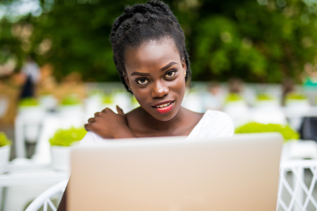 Beautiful young black female entrepreneur is sitting in street bar and working remotely on her project with laptop.の写真素材