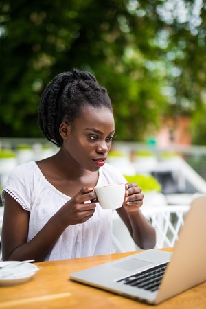 Portrait of african woman using mobile phone at an outdoor cafe with laptop and cup of coffee on tableの写真素材