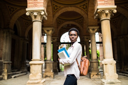Portrait of a cheerful smiling african student girl wearing backpack and holding books at universityの写真素材