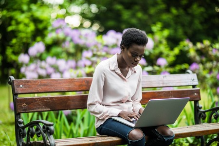 African-American female with the laptop is sitting on the wooden bench. cheerful cute black girl on the park bench using the netbook.の写真素材