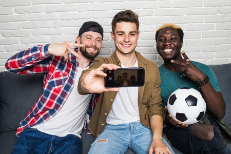Attractive guys are drinking beer, doing selfie using smart phone and smiling while sitting on couch at homeの写真素材