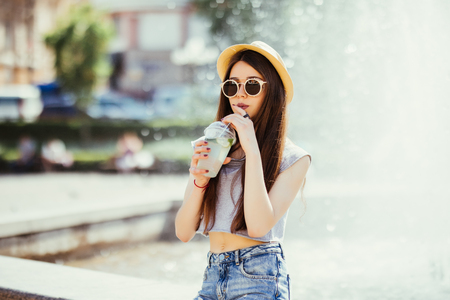 Summer sunny lifestyle fashion portrait of young stylish hipster woman walking on the street, wearing cute trendy outfit, drinking tasty smoothie, smiling enjoy her weekends, travel with backpackの写真素材