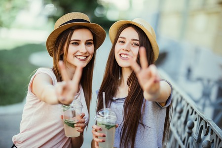 Two joyful girls taking a selfie while walking todether with cocktails on the street and showing...