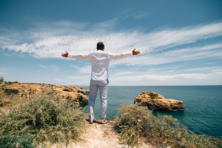 Young tourist man standing on the edge of reef enjoying ocean or sea view of turquoise water with raised handsの写真素材