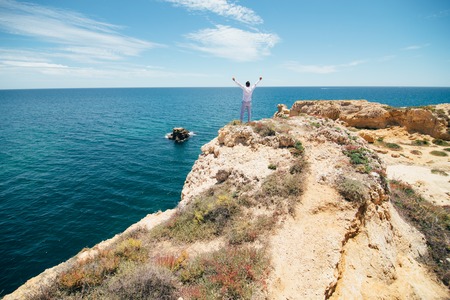 Young handsome man with raised hands at the edge of cliff enjoy the freedom view of oceanの写真素材