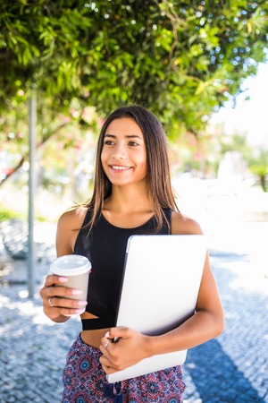 Young beautiful latin student with laptop and cup of coffee studying in the park. Girl is walking in...