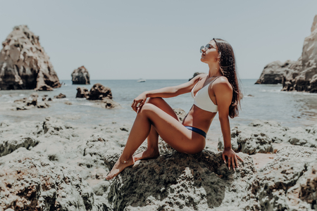 Portrait of young beautiful girl in white bikini sitting at rocks near blue green clear ocean sea water. Sunbathing woman.の写真素材