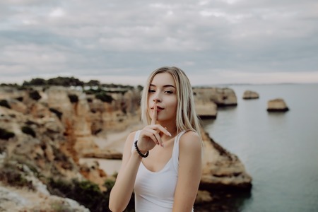 Portrait of attractive woman with silence gesture posing on rocky beach with beauty ocean landscape.