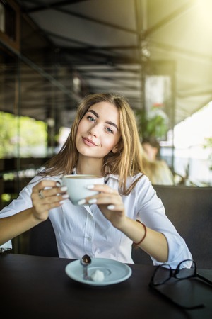 Portrait of happy young business woman with mug in hands drinking coffee in morning at restaurant