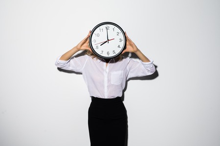 Image of young business woman standing isolated over white background covering face with clock.