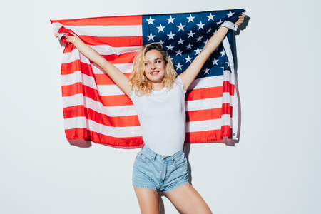 Happy young woman holding American flag and looking at camera while standing against white backgroundの写真素材