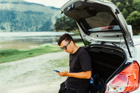 Young travel man sitting in car trunk typing on phone with beauty nature landscape on backgroundの写真素材