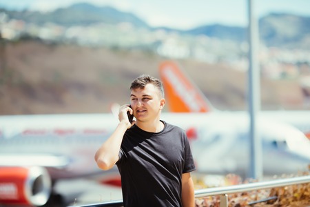 Young casual man standing on airport lounge balcony and talking on mobile phone with plane on background.の写真素材