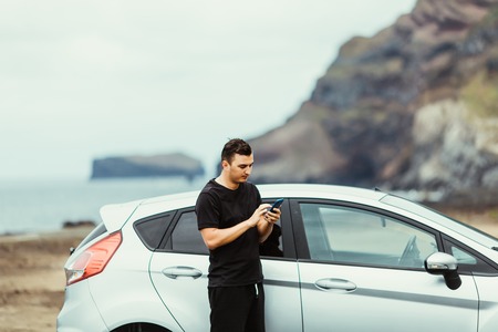 Young man driver near car use phone on summer vocation with ocean rock on backgroundの写真素材
