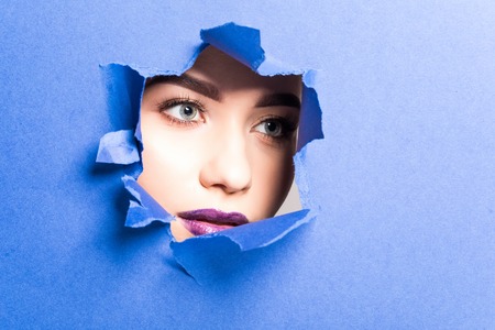 The face of a young beautiful girl with a bright make-up and puffy purple lips peers into a hole in violet paper.の写真素材