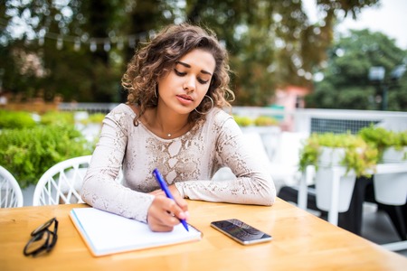 Latin female student writing in copybook at sidewalk cafe. Portrait of Caucasian woman wearing white T-shirt sitting at table by university cafeの写真素材