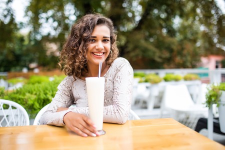 Young beautiful woman with curly hair drinks milkshake in summer in a park in the city and laughs.の写真素材
