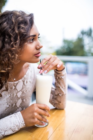 Young beautiful woman with curly hair drinks milkshake in summer in a park in the city and laughs.の写真素材