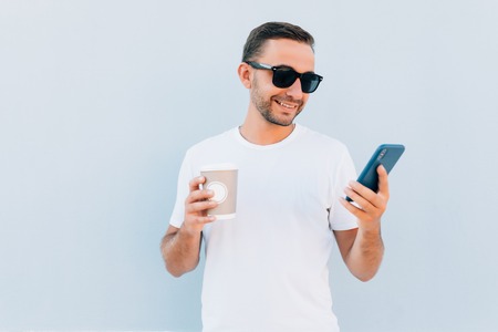 Happy adult guy in sunglasses is looking at screen of mobile phone isolated on blue backgroundの写真素材
