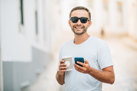 Coffee break on a go. Low angle view of confident young man in glasses holding coffee cup and using his smart phone while walking outdoorsの写真素材