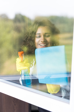 Young woman in yellow gloves cleaning window with blue rag and spray detergent. Spring cleanup,...