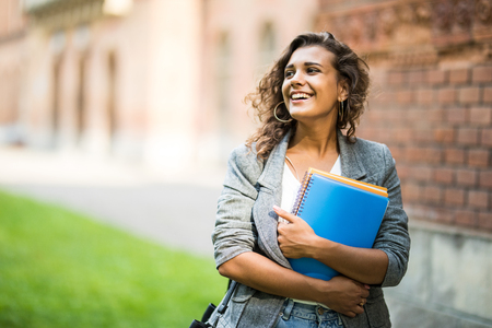 Pretty cheerful student smiling at camera carrying notebook on campus at collegeの写真素材