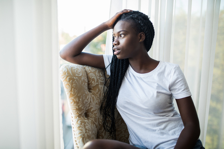 Young african american girl sitting on the chair and looking out of the window.の写真素材