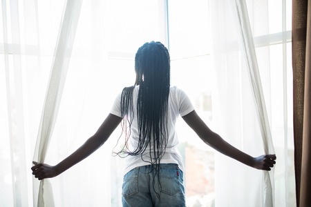 Back view of stretching african woman with curly hair in bed near window in the morningの写真素材