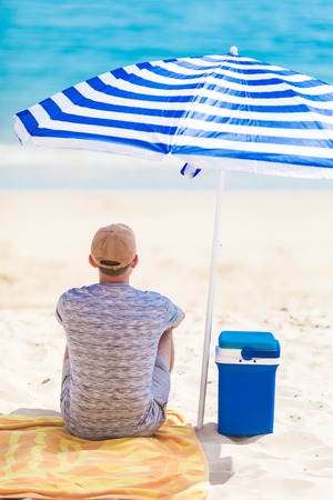 Back view of tourist sitting on the beach under sun umbrella near cooler with cold drinks.の写真素材