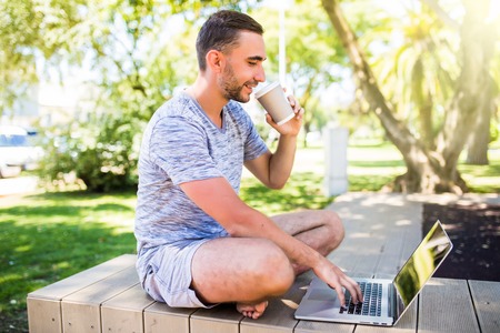 Portrait of young man with laptop and cup of coffee on bench in parkの写真素材