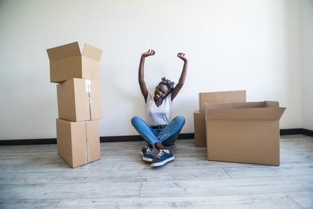 people, moving new place and repair concept - happy african american young woman with many cardboard boxes sitting on floor at homeの写真素材