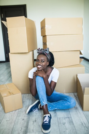 Charming young african girl standing on her knees on the floor and smiling at the camera while taking plates out of the boxの写真素材