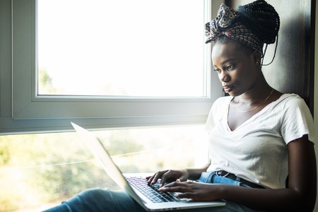 Beautiful young african woman sitting on the windowsill with a laptop.