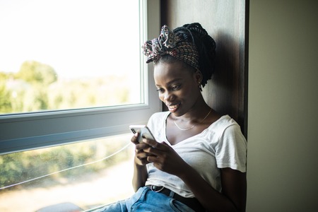 Smiling afro american woman sitting on the windowsill and writing message on phone at home