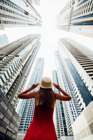 Rear view of young woman in red dress holding with both hands straw hat amazed looking up on skycrapers at downtown in modern city.の写真素材