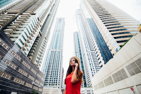 Young woman in red dress and straw hat talking on the phone in front of skycrapers in downtown of modern city.の写真素材