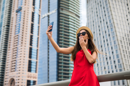 Portrait of young woman in red dress, sunglasses and summer hat make selfie or video call blow kiss on the phone on downtown skycrapers backgroundの写真素材